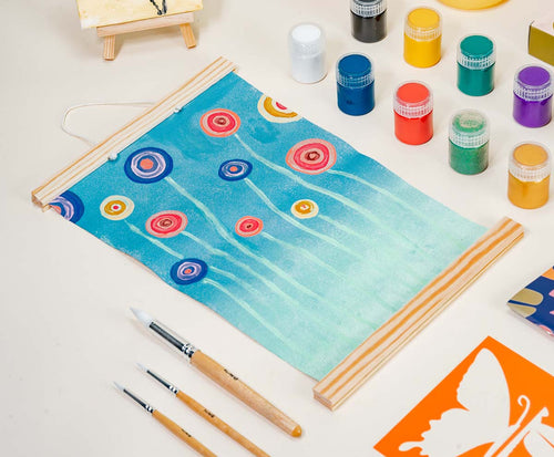 A handmade hanging scroll painting with abstract circular flowers, surrounded by paint supplies made with Sculpd Kids Canvas Painting Kit